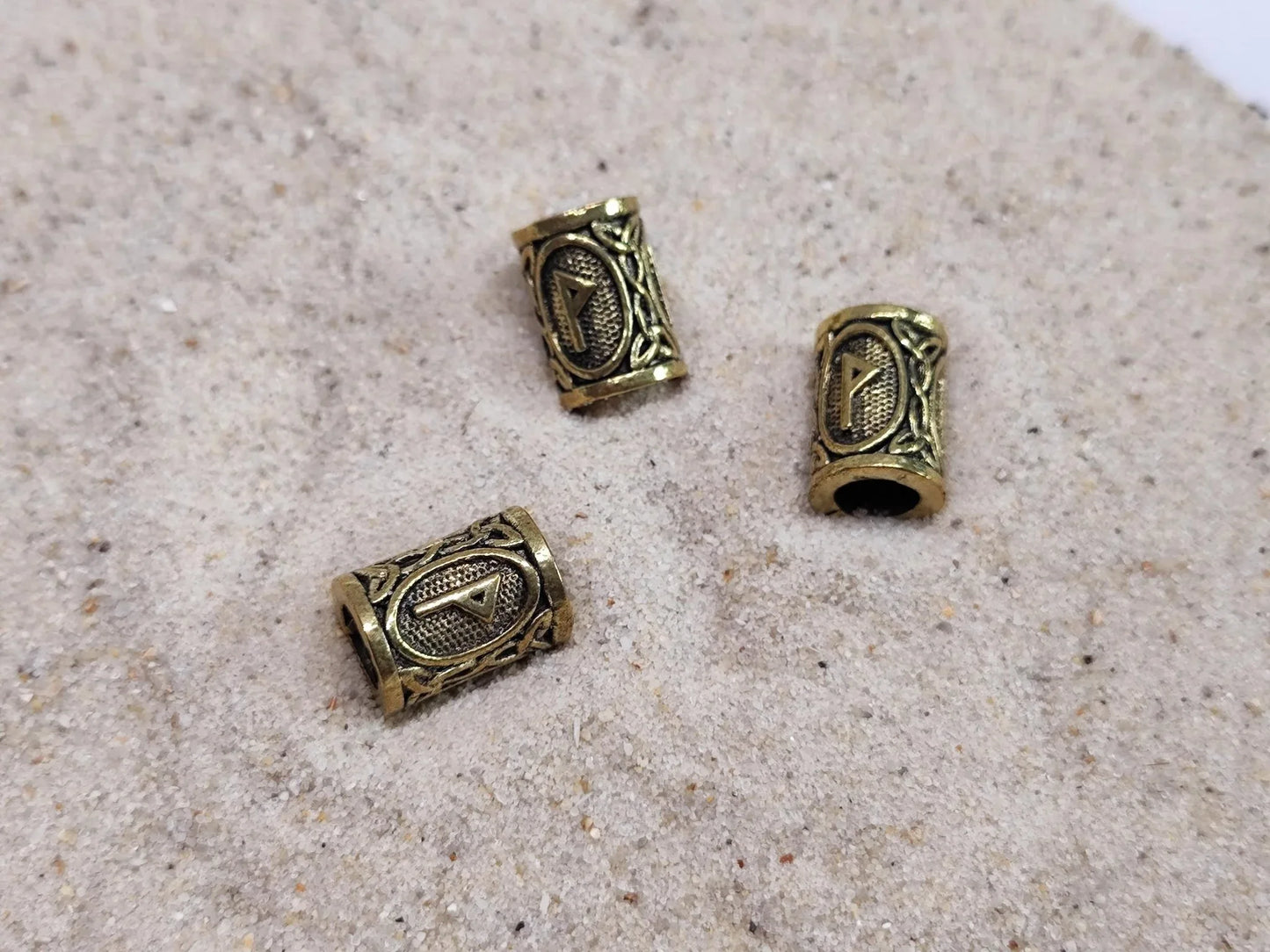 Zwei goldene Dreadperlen mit Runen im Wikingerstil auf Sand.