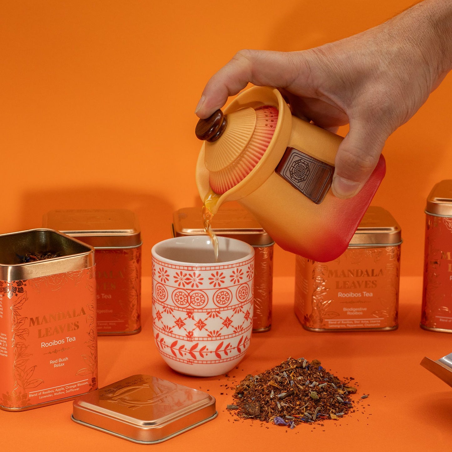 A hand pours amber tea from a yellow and red teapot into a white mug with red mandala patterns.