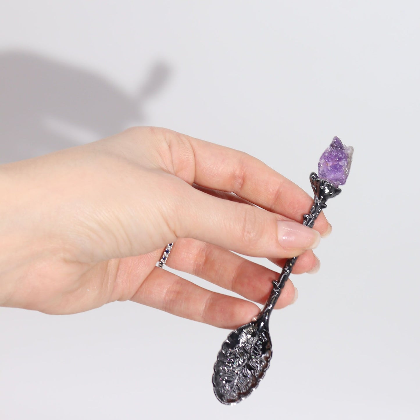 A hand holds a dark silver spoon with a purple crystal at the tip.
