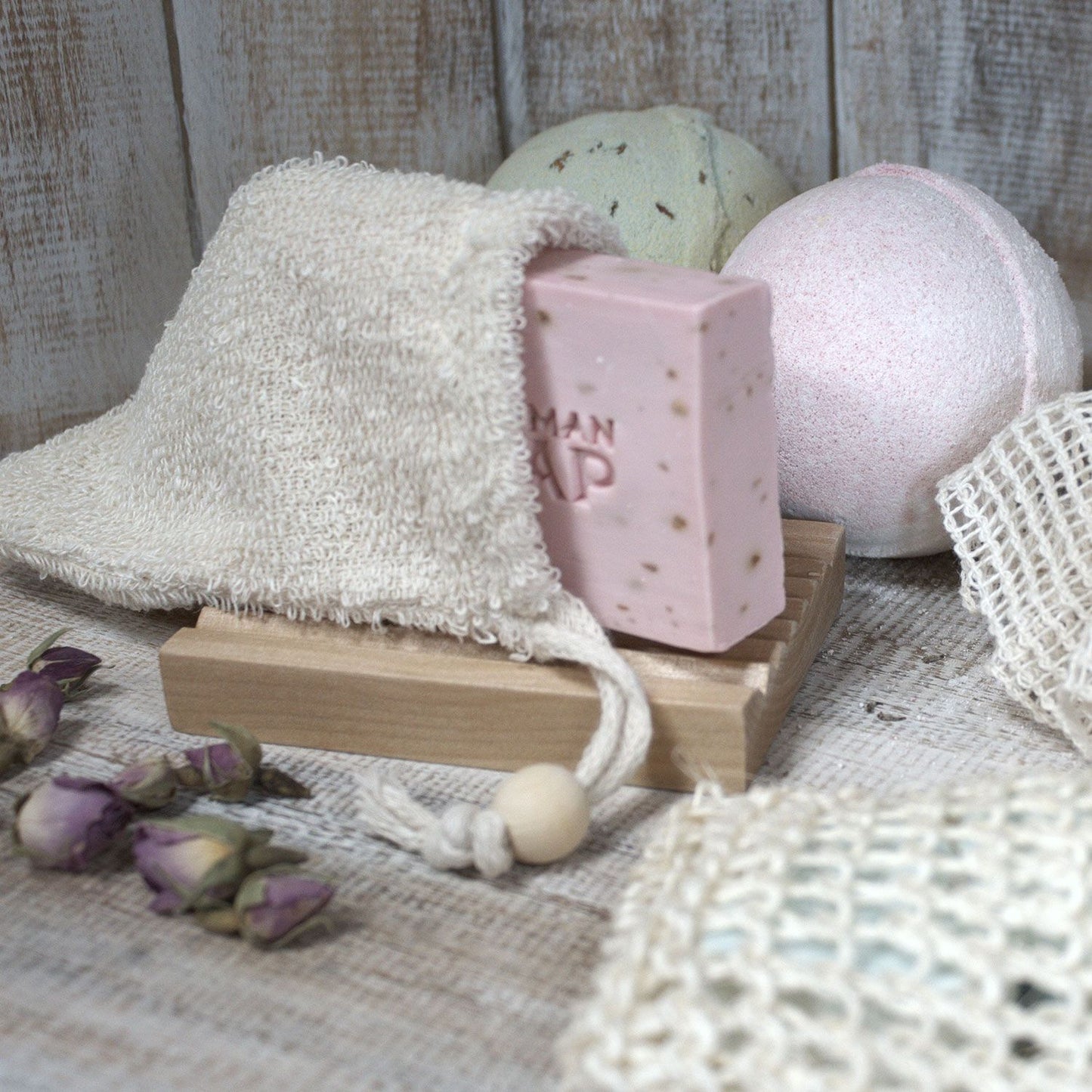 A pink bar of soap rests in a jute bag next to bath bombs and dried roses.
