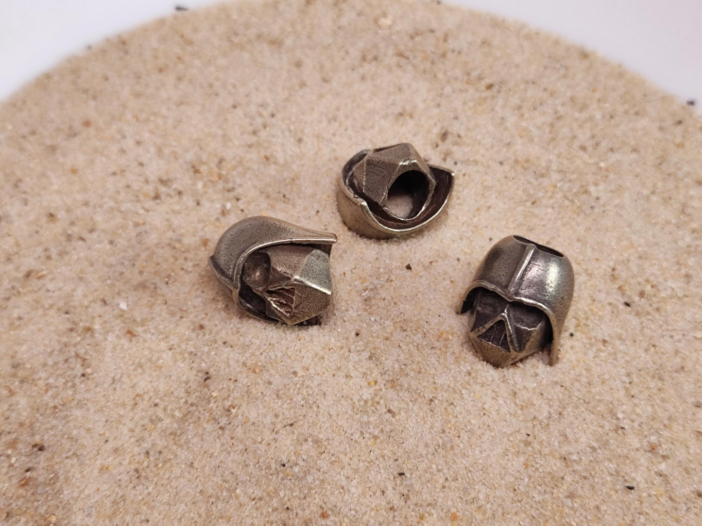 Drei Darth Vader Perlen aus Messing liegen im Sand.