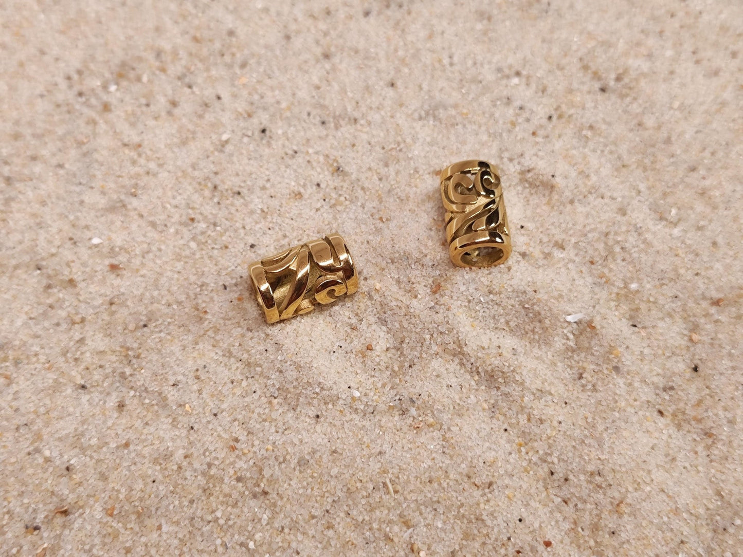 Zwei goldene Edelstahl-Dreadperlen mit filigranem Muster auf Sand.