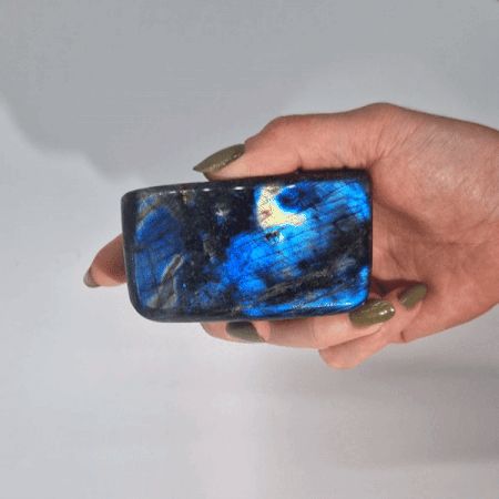 Ein rechteckiger Labradorit mit blauem und schwarzem Farbspiel wird in einer Hand gehalten.