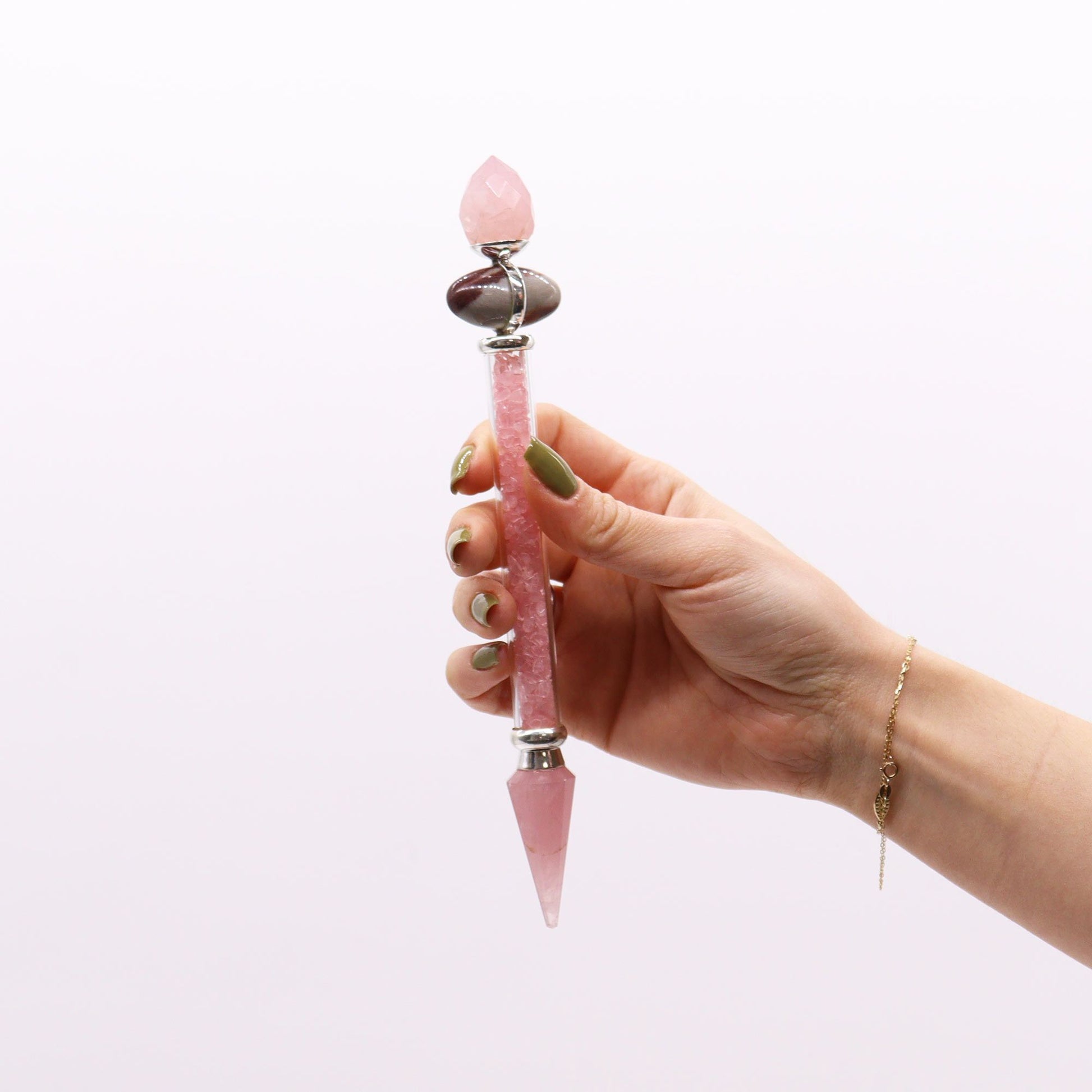 A hand holds a rose quartz and shiva lingam wand with a gold bracelet.