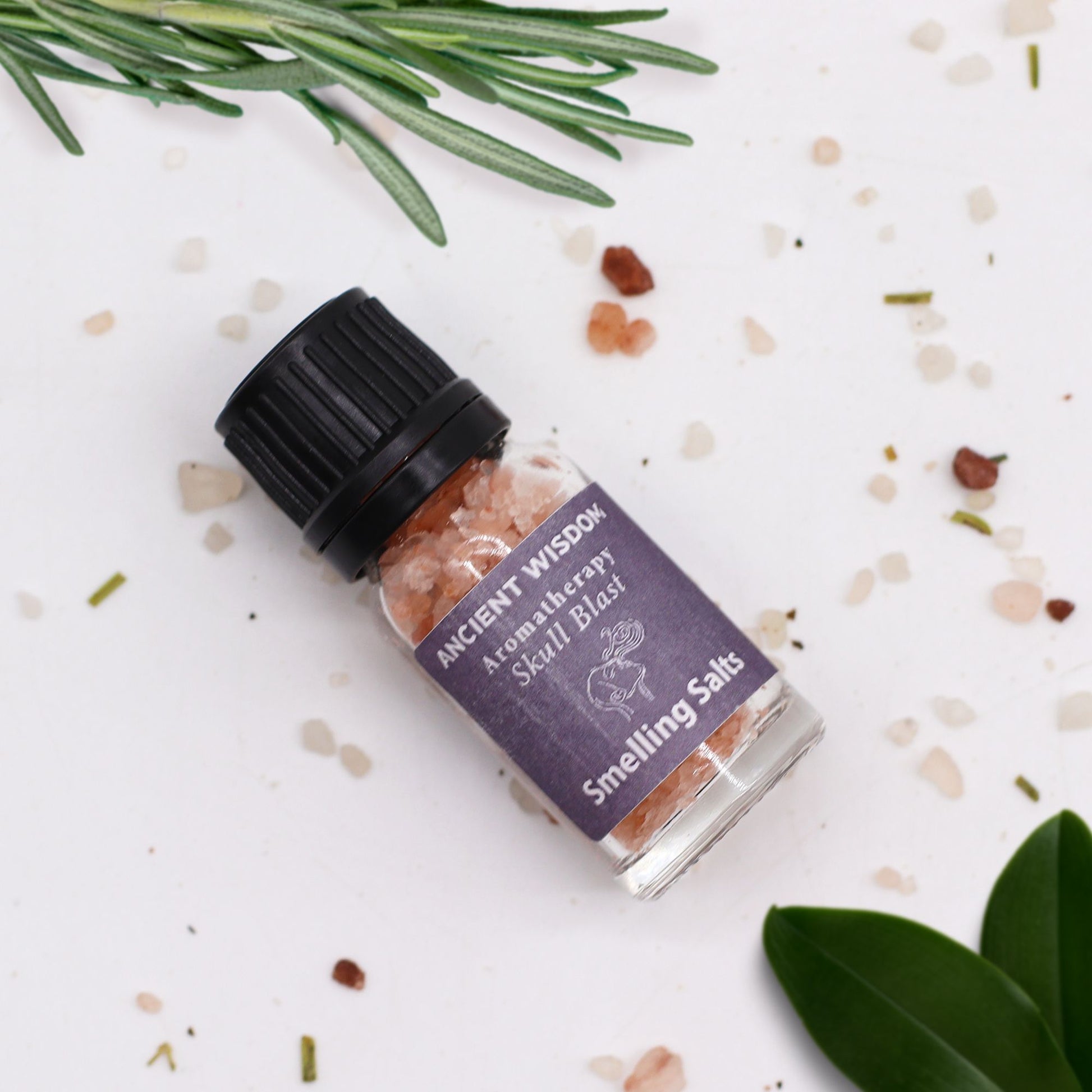 A small glass bottle of pink smelling salts with a black cap, labeled "Ancient Wisdom Aromatherapy Skull Blast Smelling Salts".