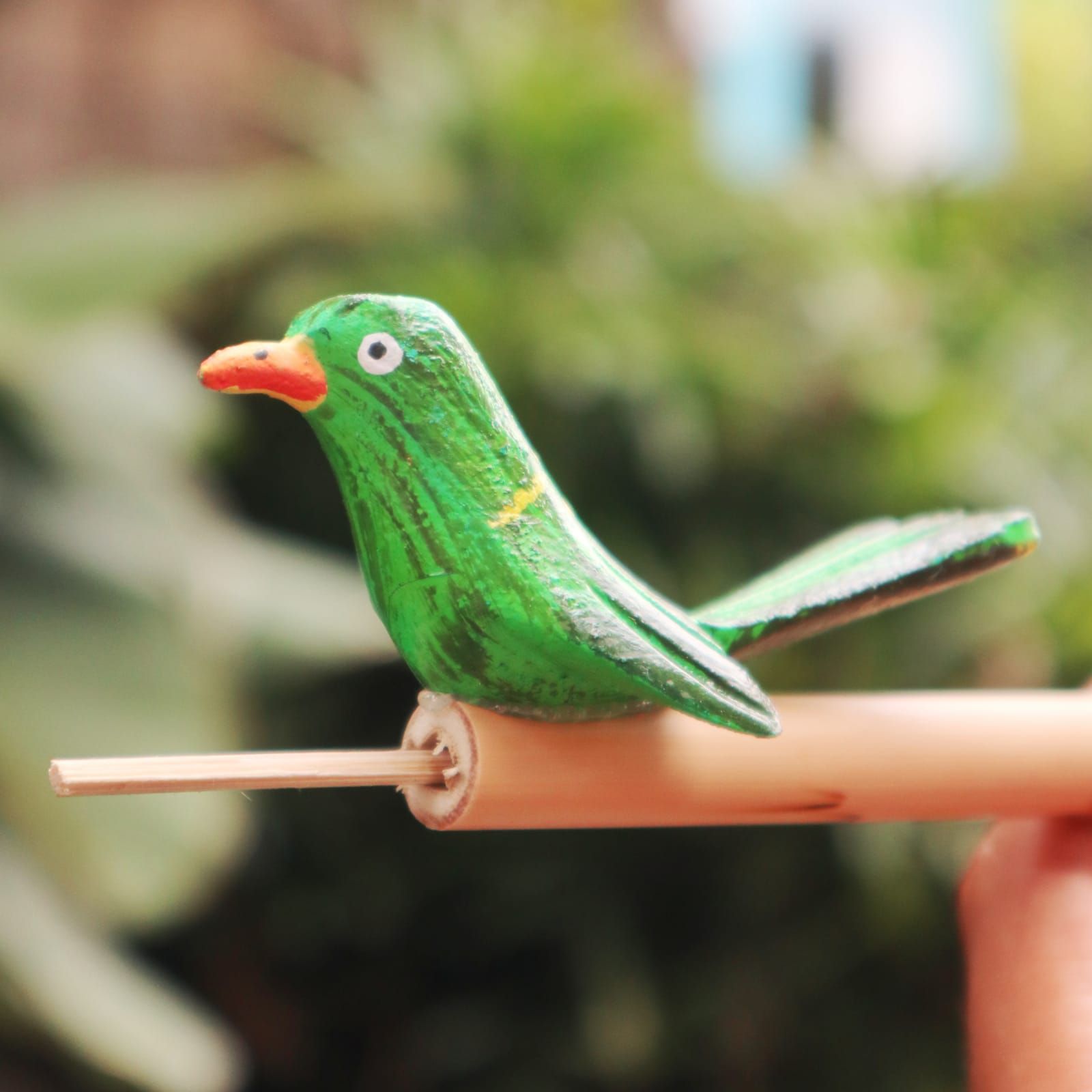 A green wooden bird whistle with an orange beak sits on a bamboo stick.