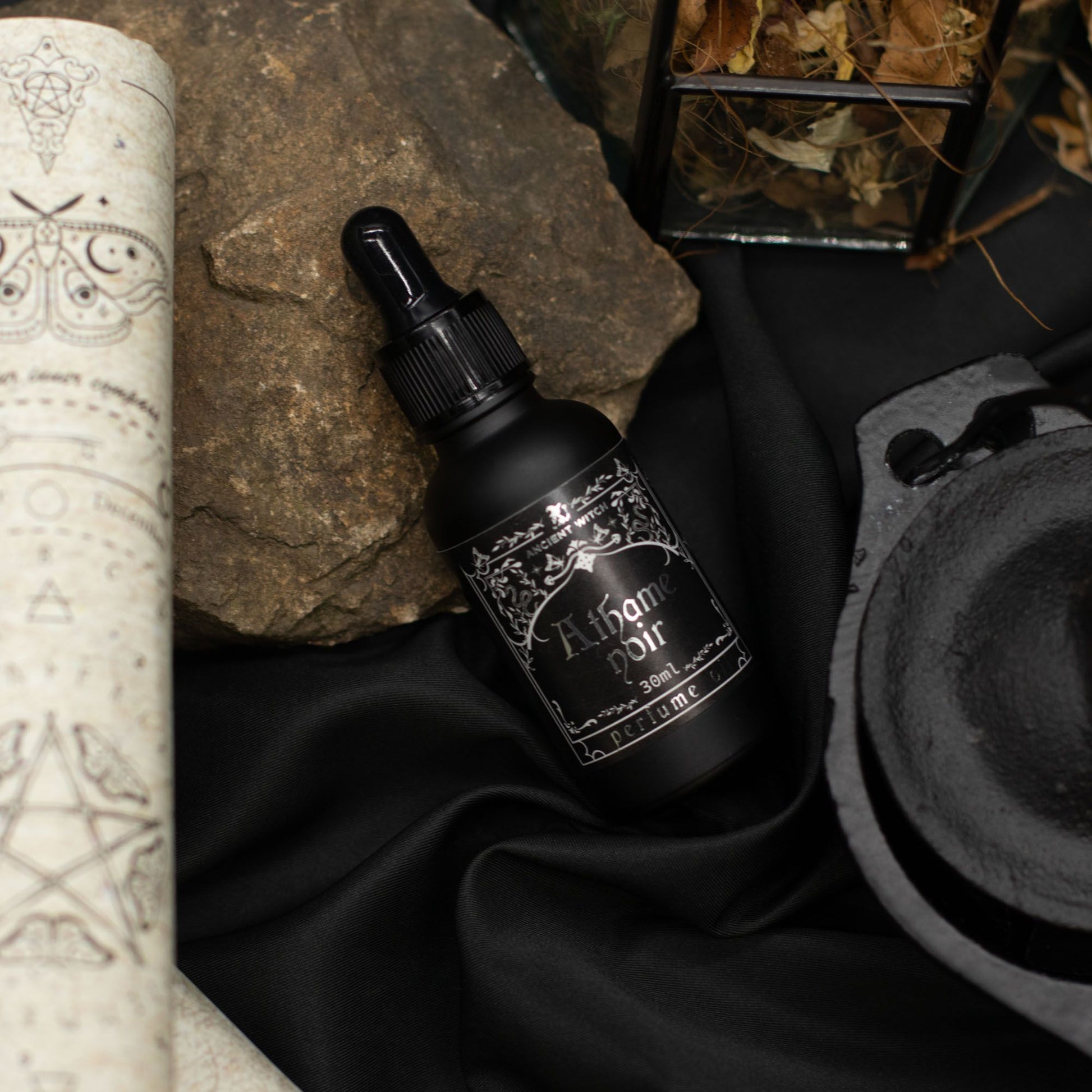 Black perfume oil bottle with Athame Noir label, on black fabric with rocks.