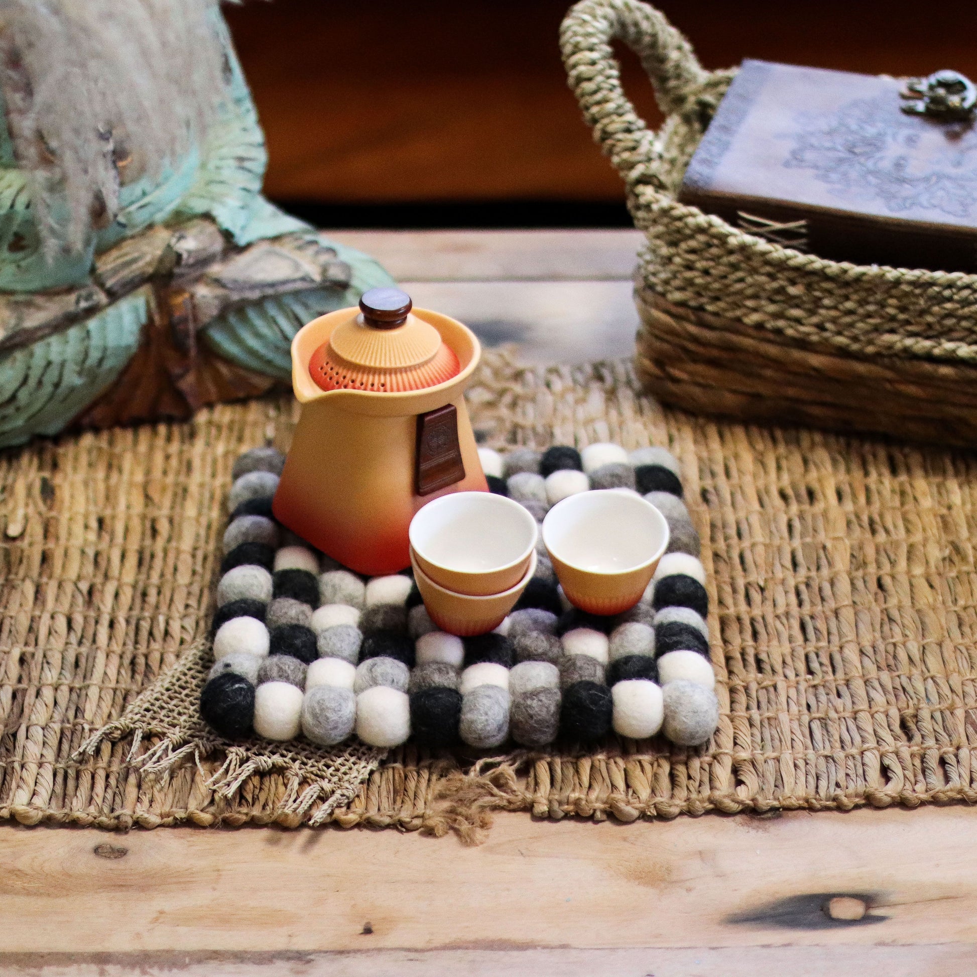 A multi-grey felt ball square placemat with a teapot and two cups.
