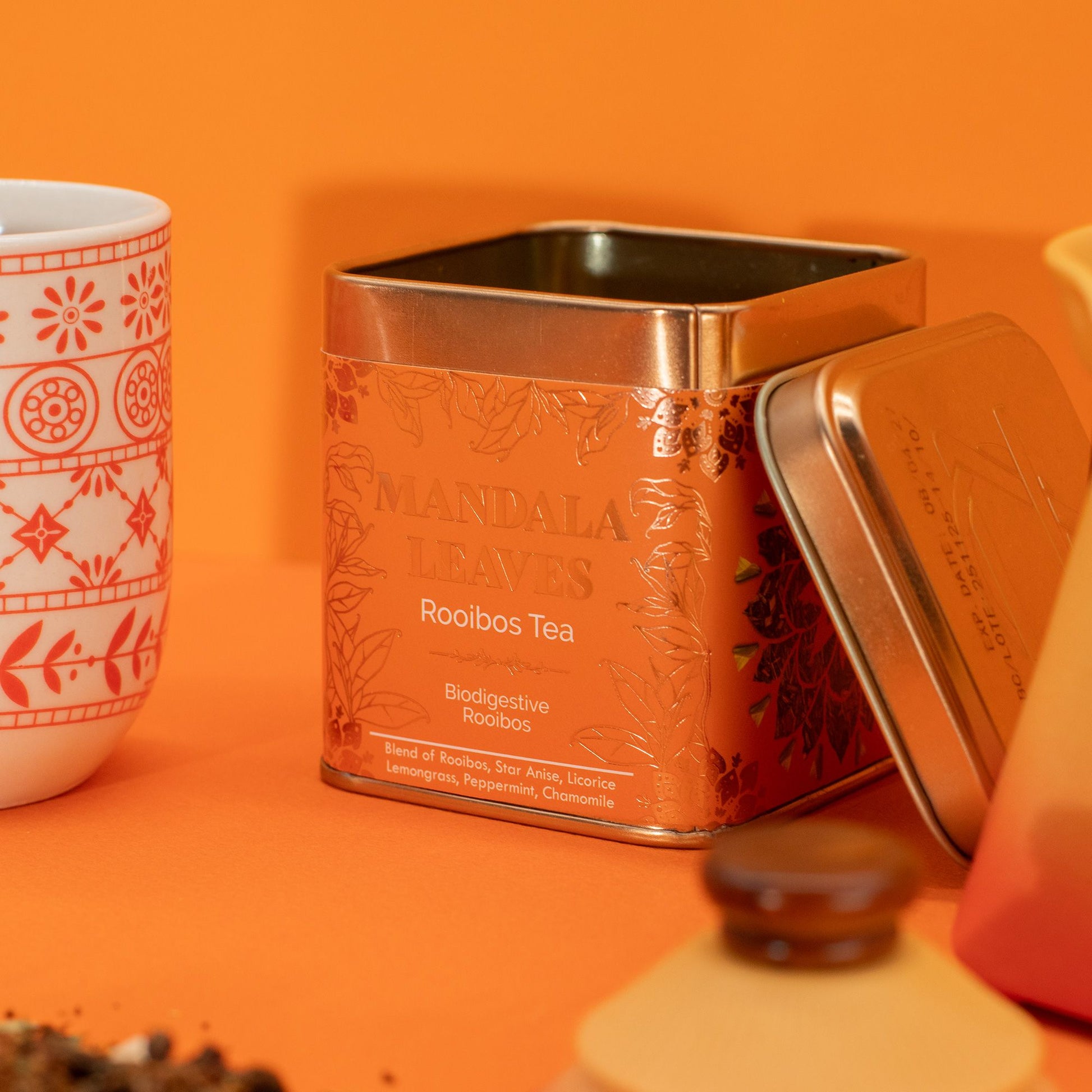 An orange tin of Mandala Leaves Rooibos Tea with a white mug.