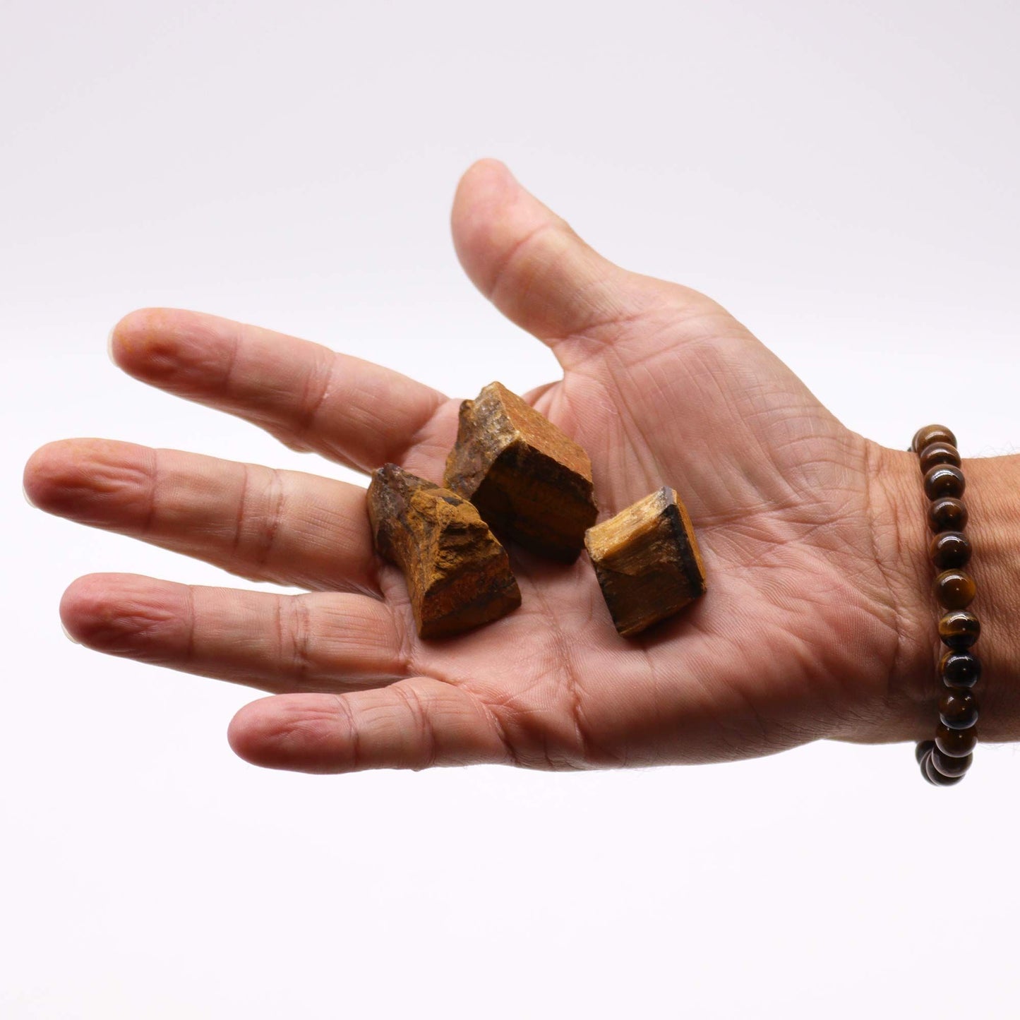 Drei rohe Tigerauge-Steine liegen in einer Hand, die mit einem Armband aus Tigerauge-Perlen geschmückt ist.