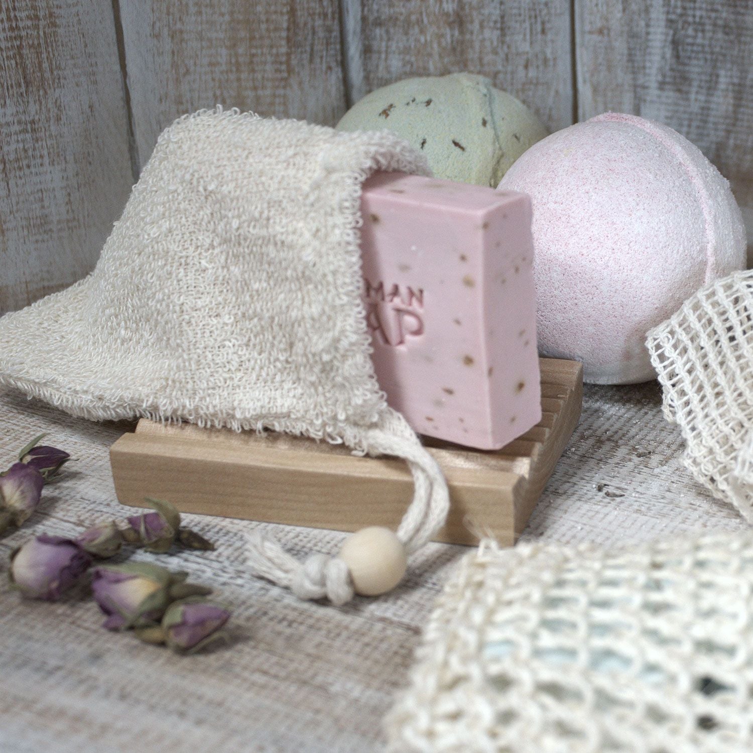 A pink bar of soap rests in a jute bag next to bath bombs and dried roses.