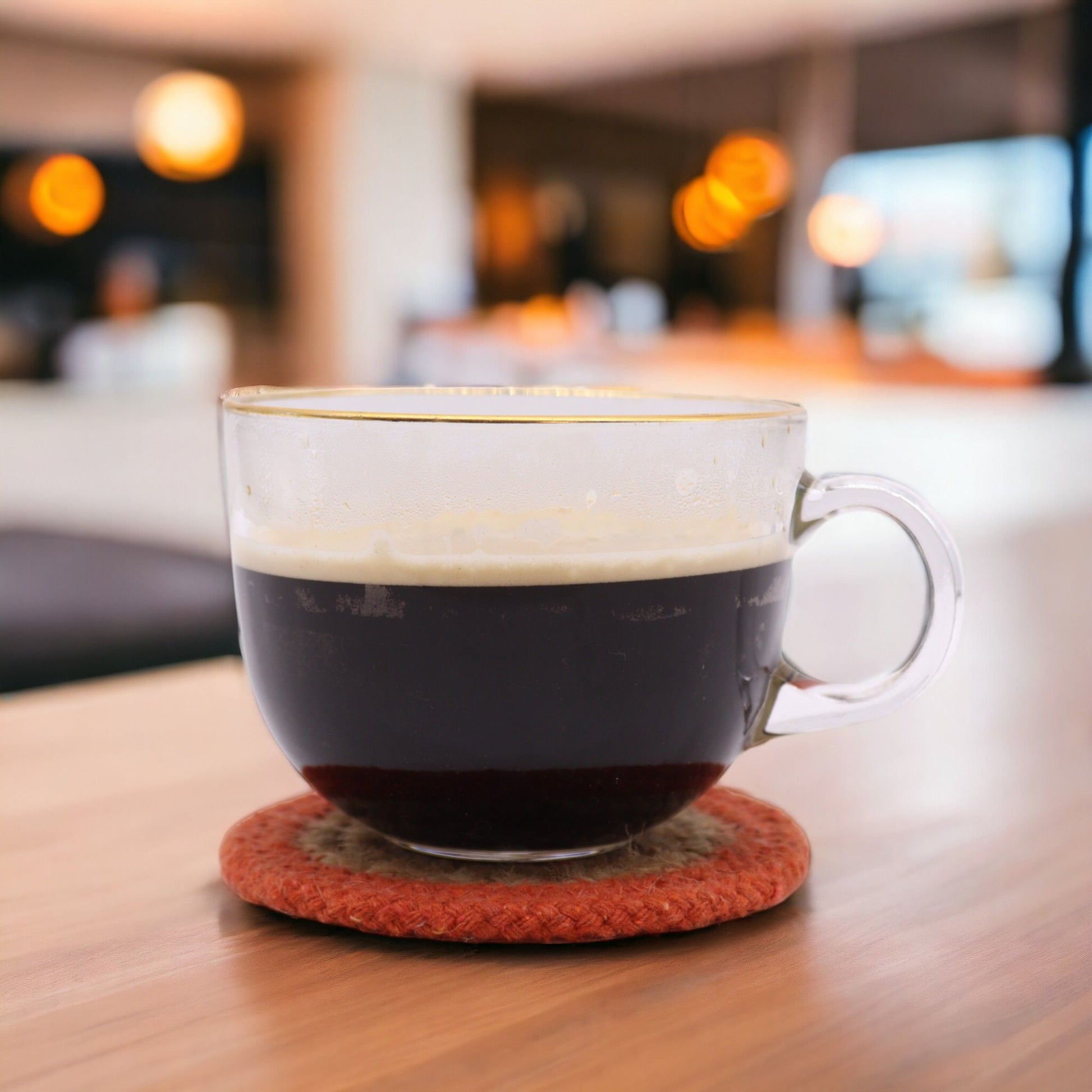 A clear glass mug of dark coffee with a foamy top sits on a round, braided jute and cotton coaster with a clay border.