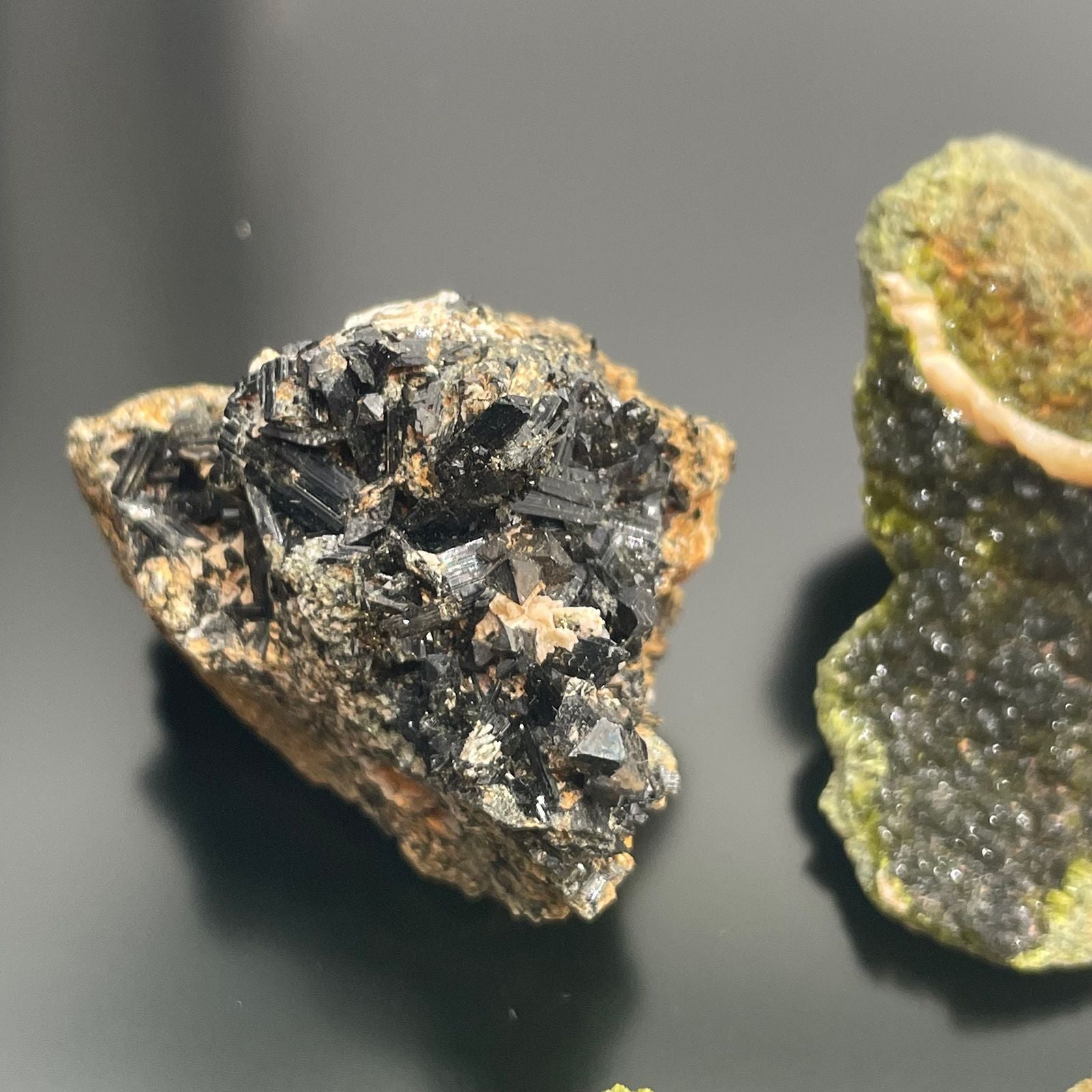Zwei rohe Epidot-Kristalle, einer dunkelgrün und der andere mit schwarzen Kristallen auf braunem Gestein.