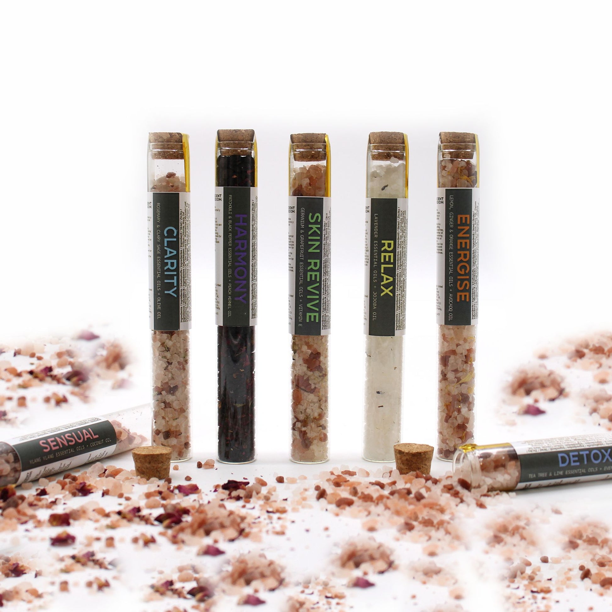 Seven vials of bath salts with cork stoppers and labels, scattered with bath salts and rose petals.
