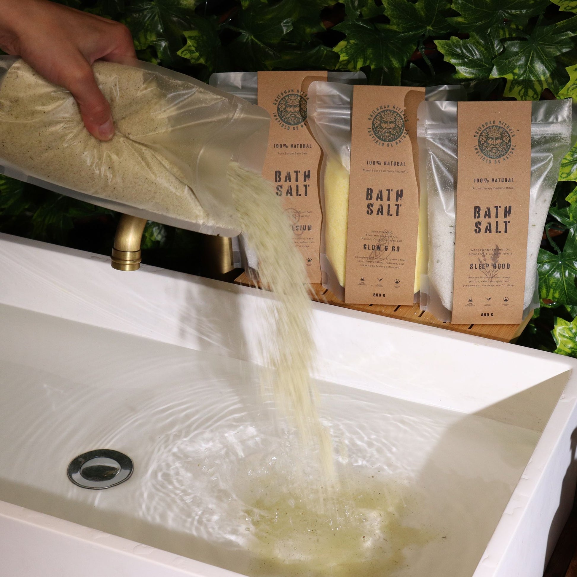 A hand pours bath salt into a bathtub with Greenman Rituals bath salt pouches in the background.