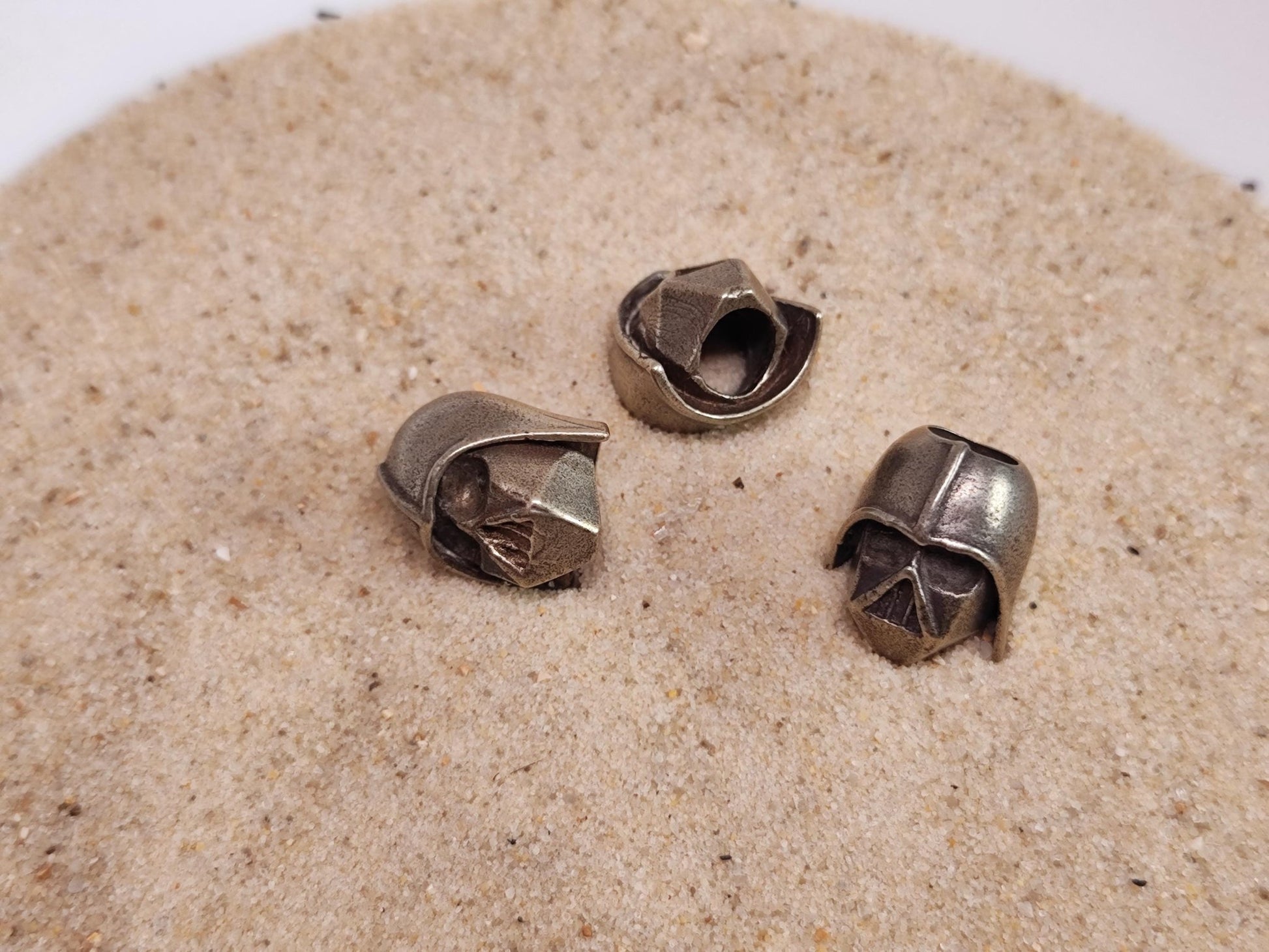 Drei Darth Vader Perlen aus Messing liegen im Sand.