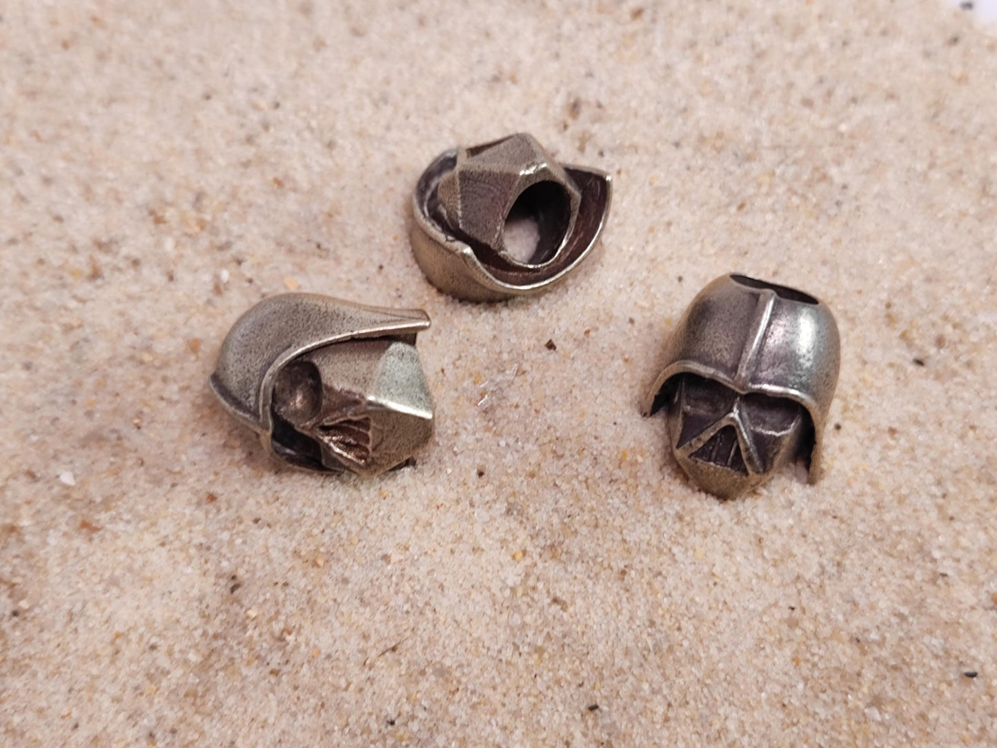 Drei Darth Vader-Helm-Perlen aus Messing liegen auf Sand.