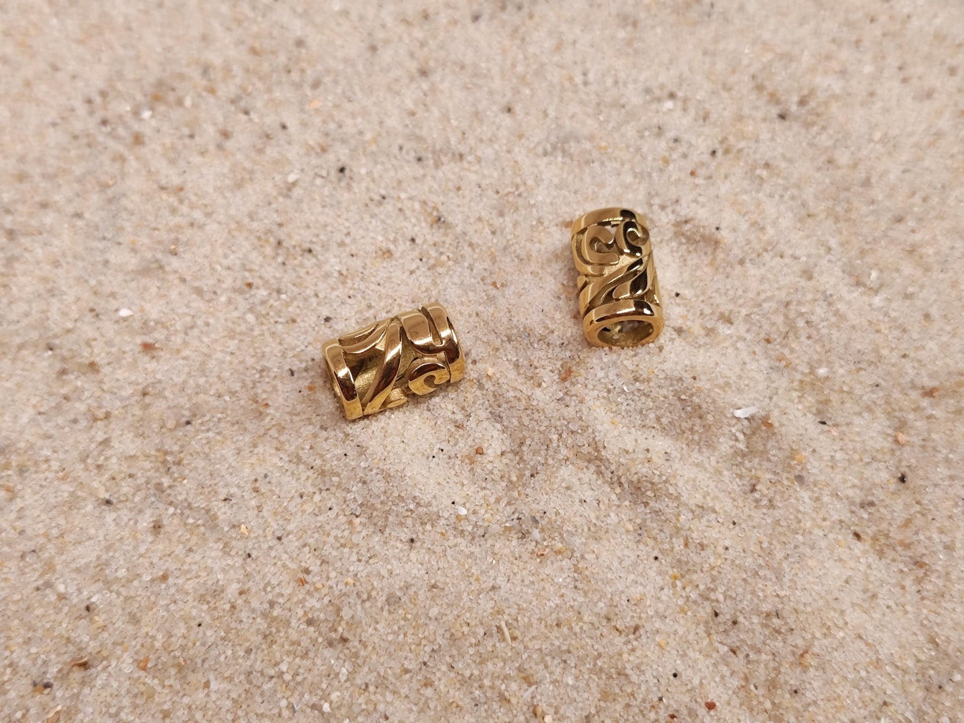 Zwei goldene Edelstahl-Dreadperlen mit filigranem Muster auf Sand.