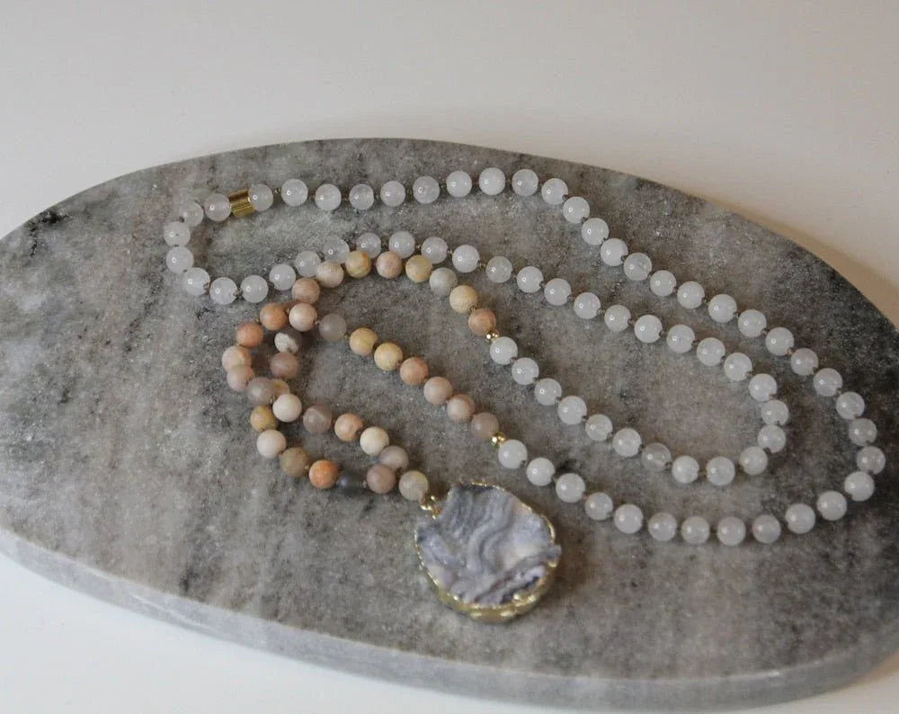 Two strands of gemstone beads, one white and one peach, with a grey agate pendant on a stone slab.